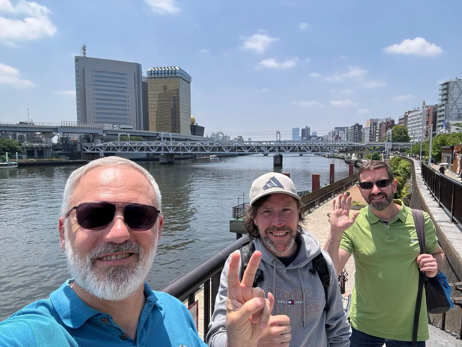 Three smiling people beside the Sumida River, with a bridge and Tokyo buildings in the background