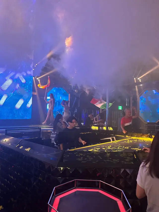 People holding flags on a club stage under blue lights and smoke, with gold confetti nearby