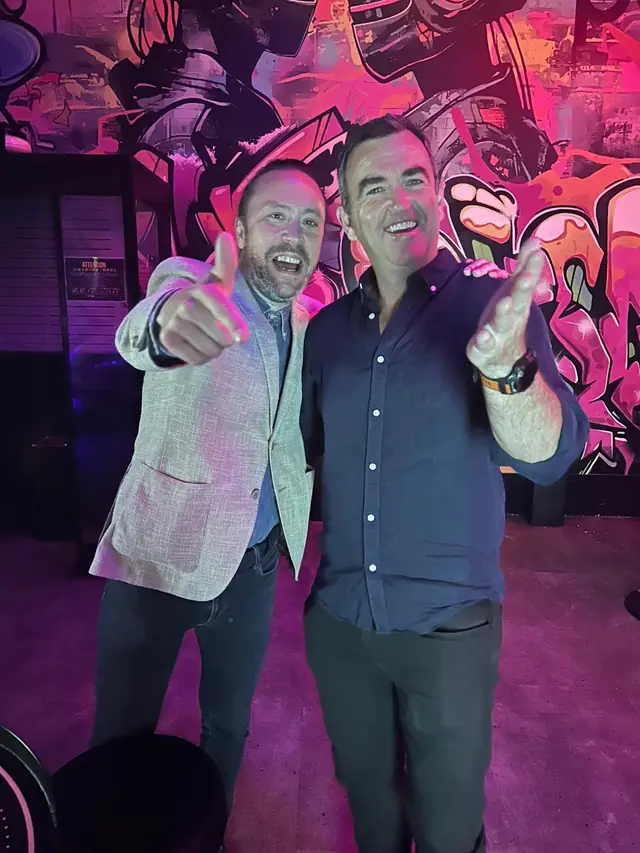 Two men smiling and gesturing towards the camera in front of a pink-lit mural