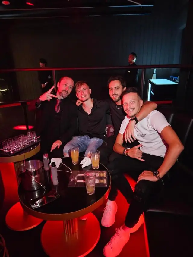 Four men seated together around a table with drinks under red club lighting