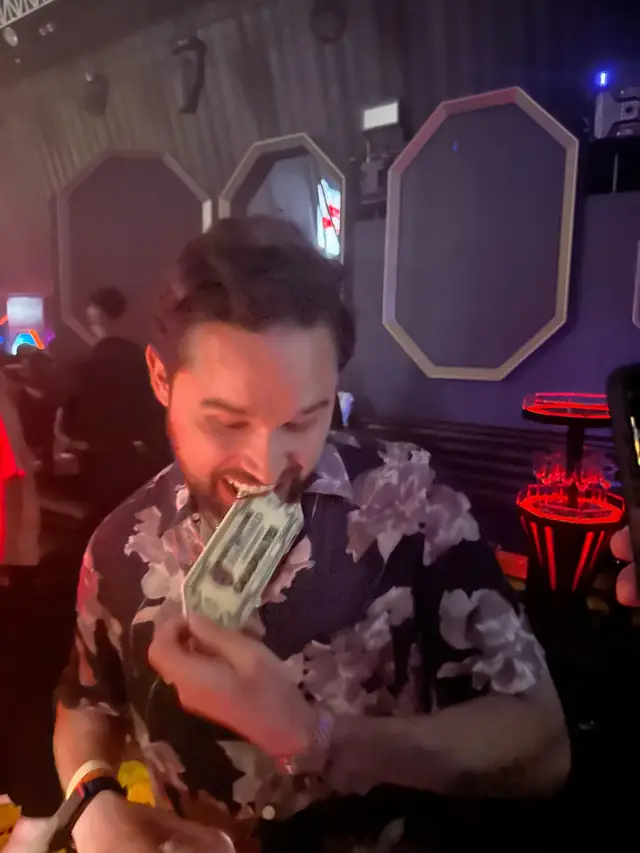 A smiling man holding a banknote-shaped object near his mouth under red club lighting