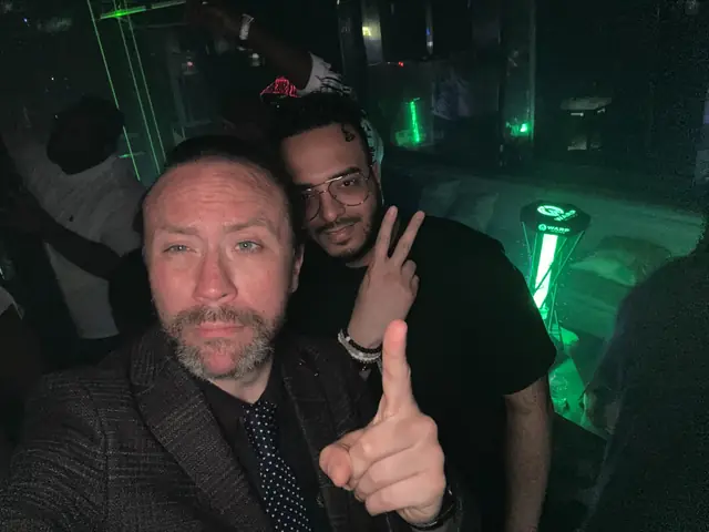 Two men taking a selfie in a club, with green lighting and an illuminated WARP logo in the background