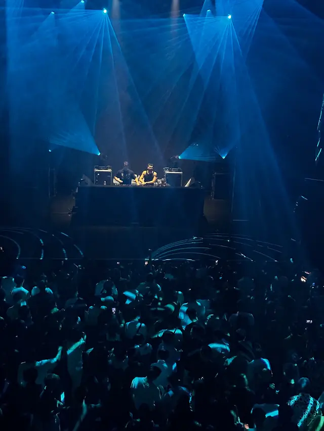 Two performers behind a DJ console above a crowd under blue beams of light