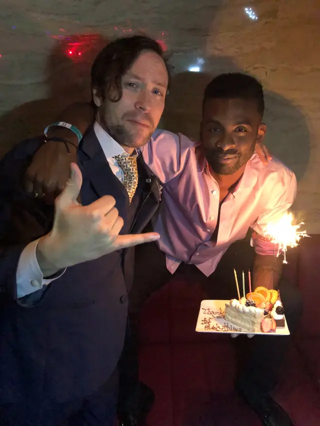 Two men posing beside a cake with fruit, candles and a glowing sparkler.