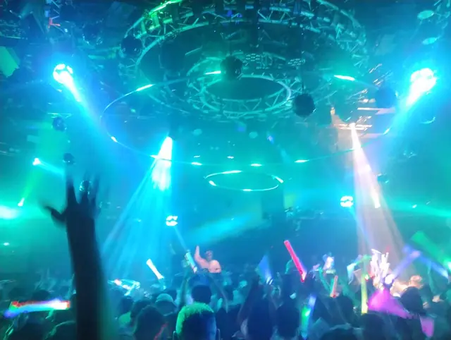 A crowd raising hands and glow sticks beneath circular blue and green club lighting