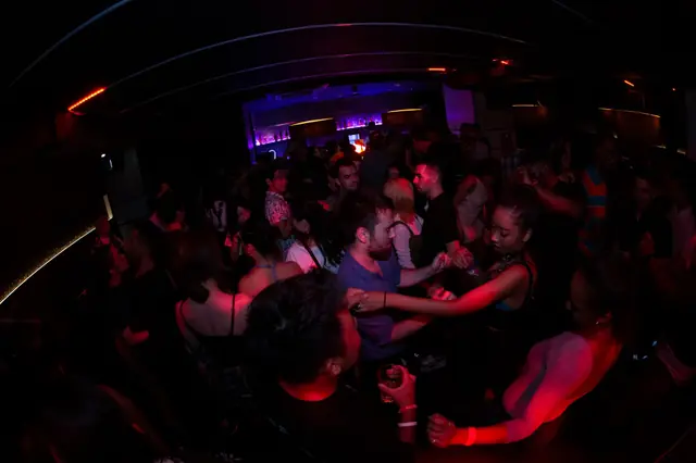 People dancing in a crowded room under red and violet lights, with a bar in the background.