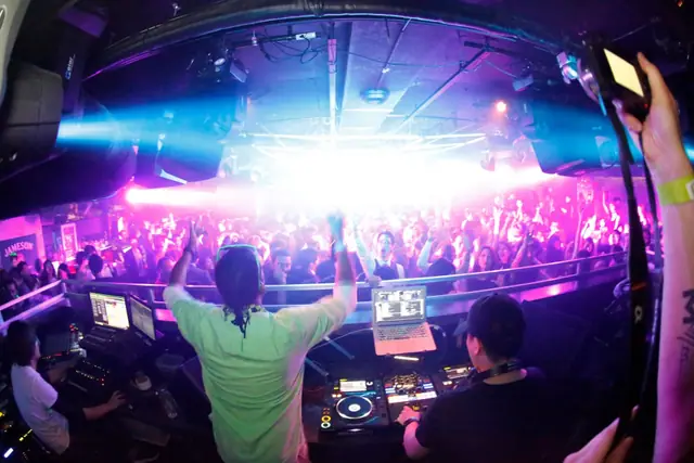 DJ booth overlooking a crowded dance floor under blue and pink lights, with people raising their arms.