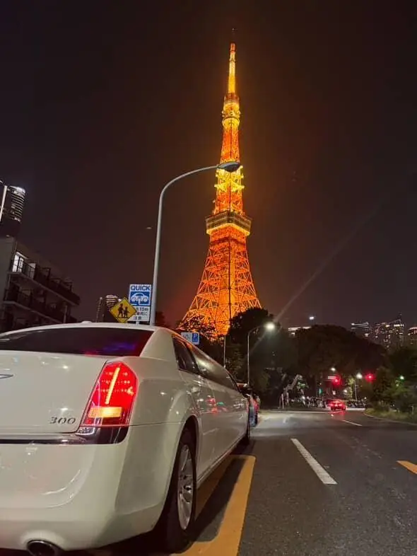 The rear of a white limousine beneath the illuminated Tokyo Tower at night.