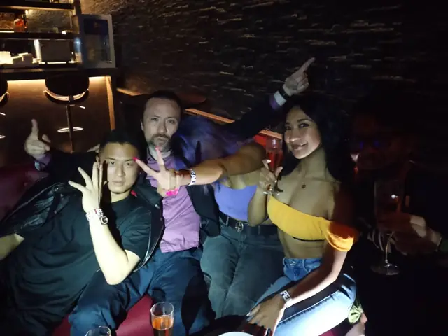 A group seated on a red lounge banquette with drinks, smiling and gesturing towards the camera