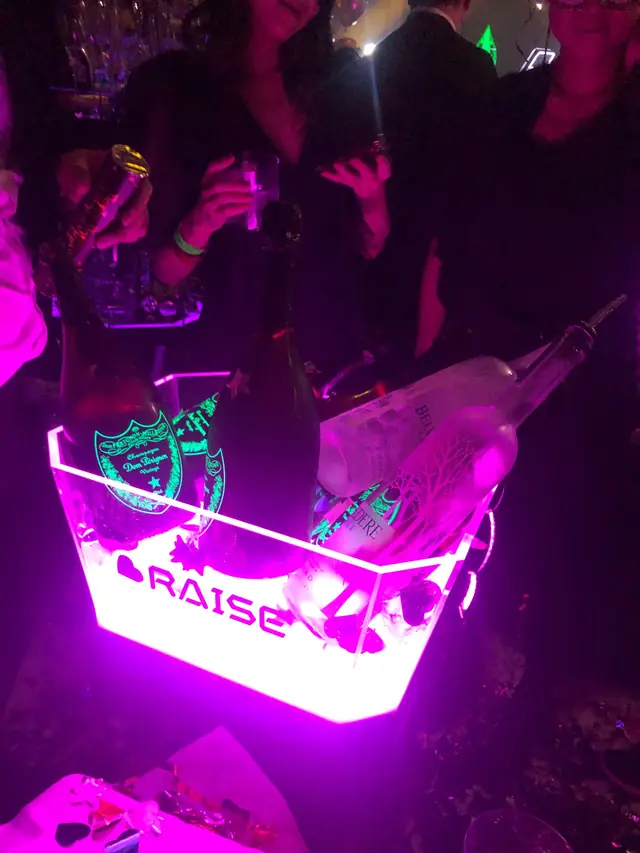 Champagne and spirit bottles in an illuminated RAISE bucket under purple club lighting