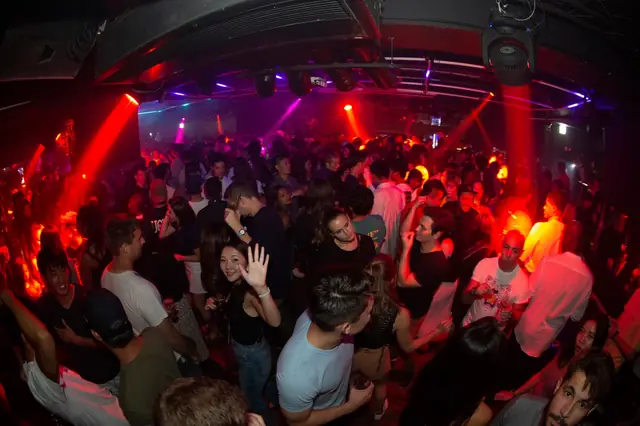Crowded club dance floor under red and purple spotlights, with a person waving towards the camera.