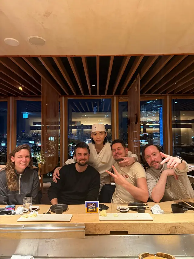 Four diners posing with a chef at a restaurant counter, with city lights through the windows